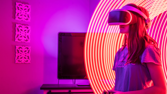 Person wearing a virtual reality headset and holding a controller indoors, illuminated by pink and purple lighting, with bright curved neon light trails forming a circular pattern around the headset. A computer monitor sits on a desk in the background.