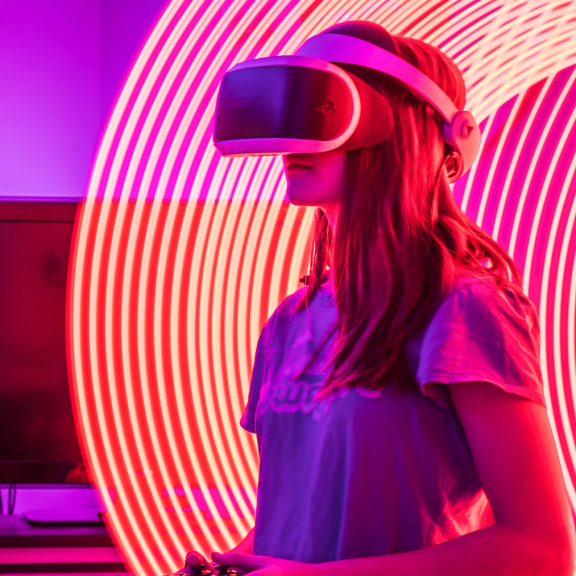 Person wearing a virtual reality headset and holding a controller indoors, illuminated by pink and purple lighting, with bright curved neon light trails forming a circular pattern around the headset. A computer monitor sits on a desk in the background.