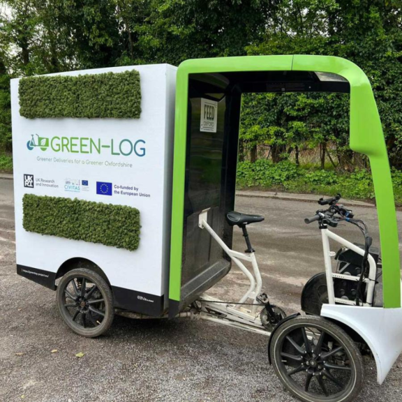 Electric cargo bike with enclosed delivery box branded “Green-Log” parked outdoors, designed for low-emission urban deliveries.