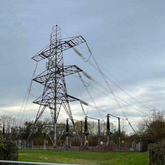 High-voltage electricity pylon and substation equipment with power lines crossing a fenced grassy area under a cloudy sky.