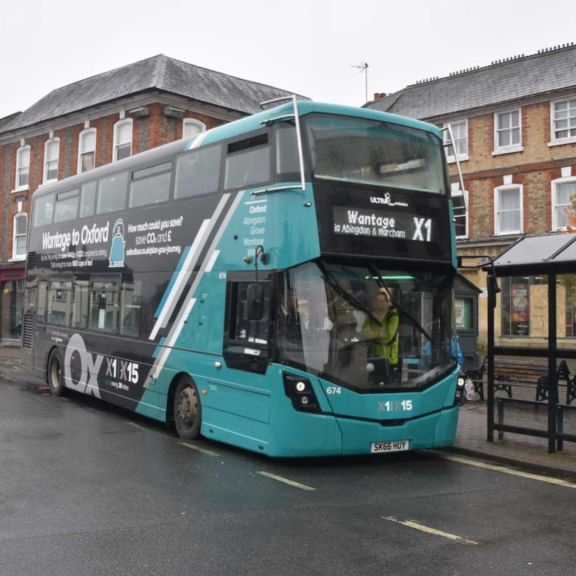 Electric double-decker bus on route X1 to Wantage, Abingdon and Oxford, stopped on a town street with nearby buildings.